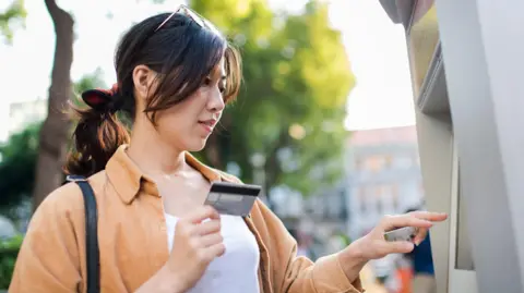Getty Images A woman uses a cash machine on the street on a sunny spring day. She holds a credit card in her hand and is pressing buttons on the machine with her other hand.