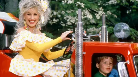 Getty Images Dolly Parton is pictured riding on a truck at her famous Dollywood theme park in Tennessee in 1977, she's in a yellow and white dresss