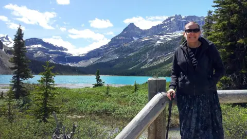 Jason S Anderson Cindy Baldassi is in the foreground wearing sunglasses and a dark hoody and navy blue dress. She is looking at the camera and behind her is a beautiful blue lake and towering mountain peaks with snow on top of them.