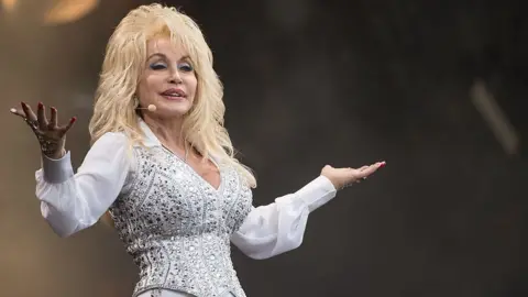 Getty Images Dolly Parton wears a white shirt and bedazzled white and silver waistcoat.