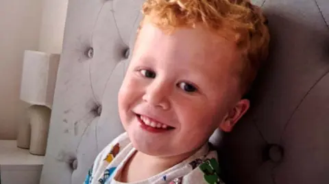 Family handout Noah, a three-year-old boy who has ginger hair. He is pictured sitting on a bed with a grey headboard and is looking directly at the camera and smiling.