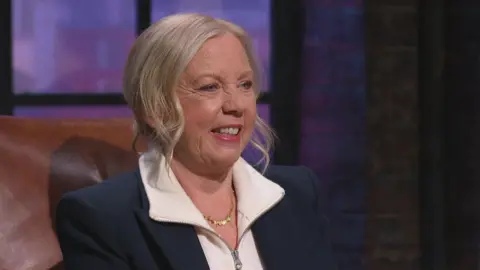 Deboarh meaden sitting in a brown leather chair wearing a navy blazer with a white, zip-up top underneath. Her blonde hair is tied back and she is smiling.