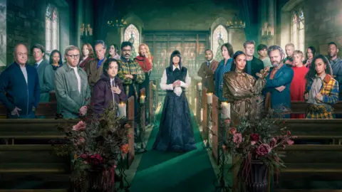 BBC The Celebrity Traitors 2 constestants all lined up inside the castle, either side of host Claudia Winkleman. They are standing in wooden pews, while Claudia stands in the aisle running down the middle, wearing a black dress with white sleeves and collar.