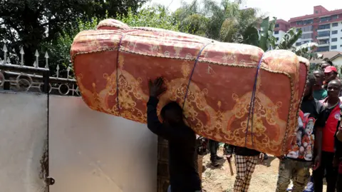 Reuters Man carrying red mattress over his shoulder. behind him is a line of people