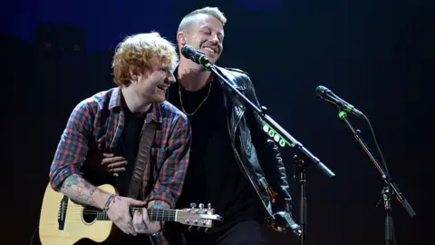 WireImage via Getty Images Sheeran holding guitar and smiling over at Macklemore who is also smiling