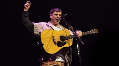 WireImage via Getty Images Rowe playing a guitar on stage in front of microphone, with one arm raised