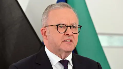 EPA/Shutterstock Australia's Prime Minister Anthony Albanese speaks at a news conference in front of several flags. He is wearing a dark suit with a red spotted tie and dark glasses