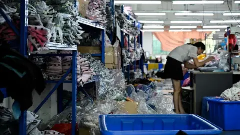 AFP via Getty Images A worker makes clothes at a garment factory that supplies Shein. She is surrounded by piles of clothes stacked on blue shelves.