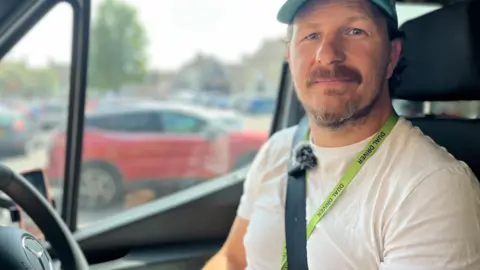 James smiles at the camera. He is sitting in the driver's seat of a minibus and is wearing a white T-shirt and a cap. He has a light green lanyard around his neck and is wearing his seatbelt. Parked cars can be seen through the window of the minibus.