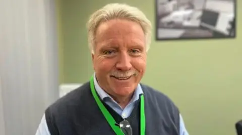 A man with blonde hair and a blonde moustache smiles into the camera. He is wearing a grey pullover with a shirt underneath and a bright green lanyard. There is a green wall behind him
