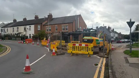 A digger sits on the edge of a corner of a junction surrounded by fencing and traffic cones