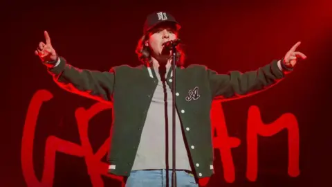 Redferns via Getty Images Singer of band Lukas Graham singing into a microphone on stage with both arms raised up
