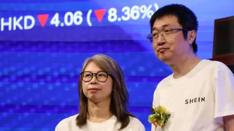 Reuters Shein's founder, Xu Yangtian (right), stands on a stage next to financial director Poppy Bao during the company's listing ceremony. Behind them is a blue screen showing Shein's shares down by 8.36%.