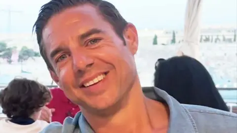 Family handout Close-up headshot of a smiling Nicholas Brandram with short dark hair, blue eyes. He is clean-shaven. He is wearing a blue open-collared shirt outdoors, with a blurred background showing an outdoor seating area and buildings under a clear sky.