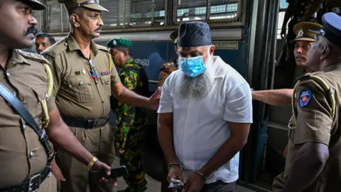 AFP via Getty Images A man with a beard, skullcap and face mask being led out of a bus by four police officers
