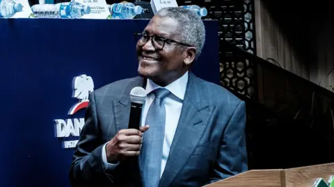 Bloomberg via Getty Images Aliko Dangote, wearing a suit, is smiling as he turns to the left of the picture. He is holding a microphone in his right hand.