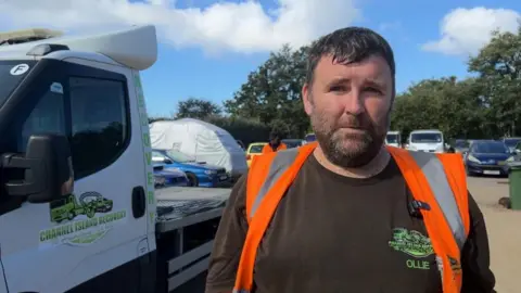 BBC A man with short dark hair and a short dark beard stands outdoors beside a white recovery truck. The person is wearing a black T-shirt and a bright orange high-visibility vest and is facing the camera. The truck door displays the logo and name "Channel Island Recovery", while several parked vehicles, a covered vehicle and trees are visible in the background under a blue sky with scattered clouds.