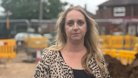 BBC A woman in her late 30s, with blonde hair, wearing a black dress and leopard print top.