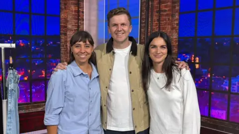 BBC Two women and a man standing in a TV studio with their arms around each other. The women have long, dark hair and the man has brown hair. They are all looking at the camera and smiling.
