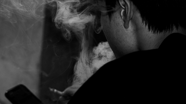 A black-and-white close-up from behind a person holding a smartphone while smoke swirls past their head.