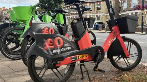 LDRS Image of a red Voi e-bike and green Lime bike parked on the side of a pavement. In the background there are rows of houses.