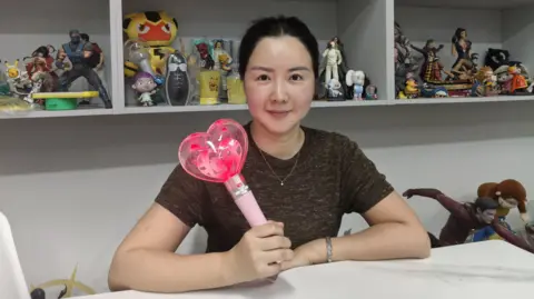 Mihox Melanie Wang from the Mihox sales team holds up a toy. She is sitting in front of shelves of other toys