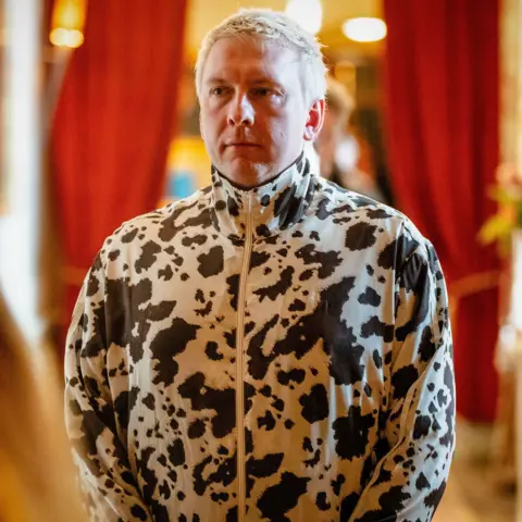 BBC/Studio Lambert/Euan Cherry Joe Lycett standing in a zipped up white jacket with numerous black spots and splodges
