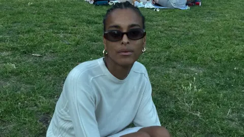 Summer Nesbeth Summer wears a white jumper and sits on the grass. She has black hair braided back and wears sunglasses and gold earrings.