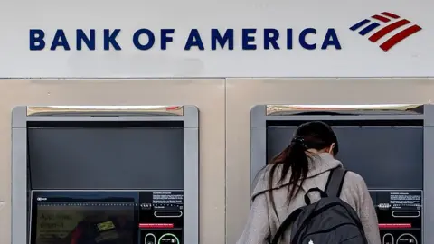 Getty Images Woman stands in front of a Bank of America cash machine. She has her back to the camera and is wearing a grey coat and black rucksack