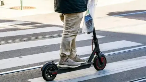 Getty Images A person wearing trainers and trousers riding a black e-scooter across a pedestrian crossing