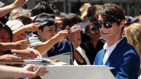 Getty Images RJ Mitte in dark glasses and a blue suit, smiling and signing autographs next to a crowd of fans holding out their arms, in 2013