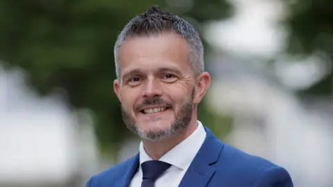 PA Media Robbie Butler. He has short grey-ish hair and beard, wearing a blue blazer, white shirt and navy tie.