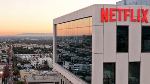 Mario Tama / Getty Images A wide view shows the Netflix logo on the side of a cream coloured building at Netflix studios in 2025 in LA, with the iconic LA skyline in the background at sunset.