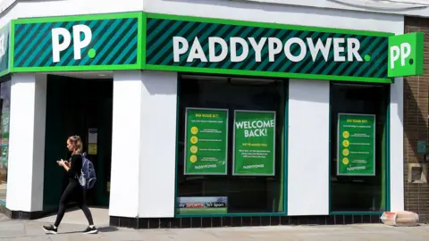 Getty Images The Paddy Power logo is seen atop a storefront of a branch in London in 2020, with a person walking in front of it along the street looking at their phone on a sunny day.