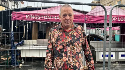 Tim Meads stands beside a security gate blocking access to a market stall while rain falls. He is wearing a shirt patterned with flowers.