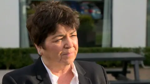 Rita Devlin has short dark hair and wears makeup and green earrings. She is wearing a black blazer and a white shirt. Behind her is a white building with a black window and curtains on the inside. There is a black bench directly behind her with a hedge next to it.