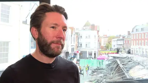 A man in a black top stands in front of a window overlooking a market area, where a fire-damaged structure with a collapsed, charred roof can be seen below.