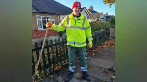 Broadhurst family Wayne Broadhurst standing on a pavement wearing hi-vis clothes and holding a broom.