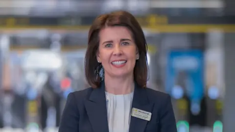 Merseyrail A woman with shoulder length hair, a grey top and a navy suit on with a badge reading her name Suzanne Grant