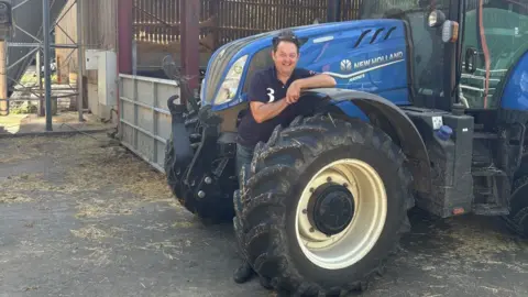 Surrey County Council Hugh Broom leans against the front wheel of a large blue tractor inside a farm building. The tractor occupies much of the right side of the image, with large tyres and agricultural equipment visible. Farm sheds, feed storage areas and metal structures can be seen in the background. Sunlight enters from the open side of the building.