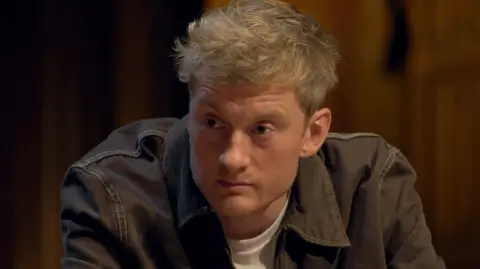 Getty Images James Acaster sitting at the round table wearing a dark denim jacket on Celebrity Traitors