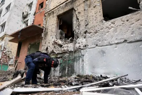 State Emergency Service Of Ukraine An official, wearing a helmet and protective gear, sifts through rubble. There is a heavily damaged building behind him.