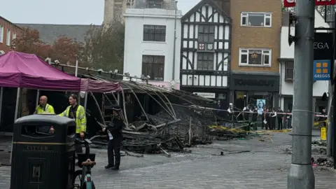 All Saints Church Charred and collapsed market stalls in Kingston upon Thames, with debris scattered across the marketplace as police officers and other people stand behind cordons. All Saints Church and surrounding shops can be seen in the background.