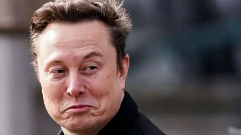 Reuters Elon Musk smiles at a camera while appearing to walk by. He is wearing a black coat and has short brown hair