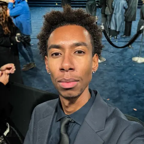 Joel Marlinarson Joel is smartly dressed in a navy suit with a black tie and poses for a selfie in front of a blue carpet. Behind him are velvet ropes for a VIP area with people waiting in the background.