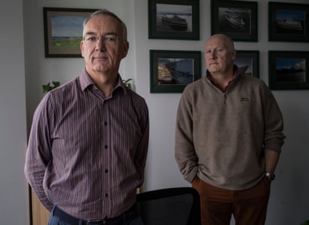 John Barton and James Bates stand indoors near walls hung with framed photographs of boats