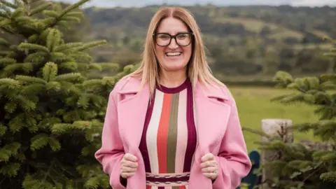 Emma Hine A woman wearing a light pink coat is standing in between two pine trees, with fields behind her.