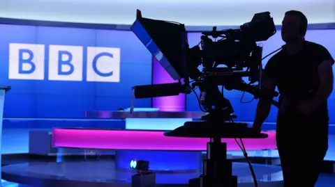 BBC A camera man seen in a studio with the BBC logo in the background against a pink and purple backdrop