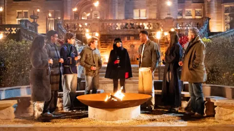 Euan Cherry/Studio Lambert/BBC/Press Association Leigh-Anne Pinnock, Romesh Ranganathan, Sebastian Croft, James Blunt, Claudia Winkleman, Kenny Oghenovo Ojuederie (aka King Kenny), Maya Jama and Michael Sheen stand around a fire pit on the set of The Celebrity Traitors. It sits in front of Ardross Castle in the Scottish Highlands, the location for the show.