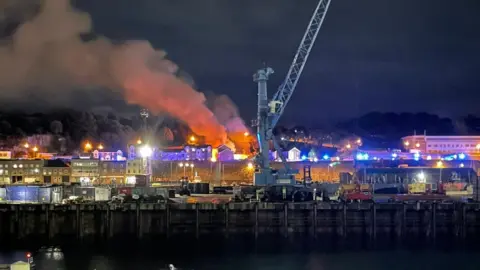 Robert Hall/BBC Plumes of smoke come out of a building behind a sea front at night. A crane and boats are in the foreground.